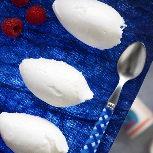 Drie bolletjes witte sorbet op een blauw oppervlak met patroon, met frambozen en een lepel.