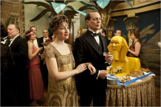 Scène d'une fête. Un couple se tient près d'une table de nourriture avec une statue de sphinx. D'autres invités en arrière-plan.