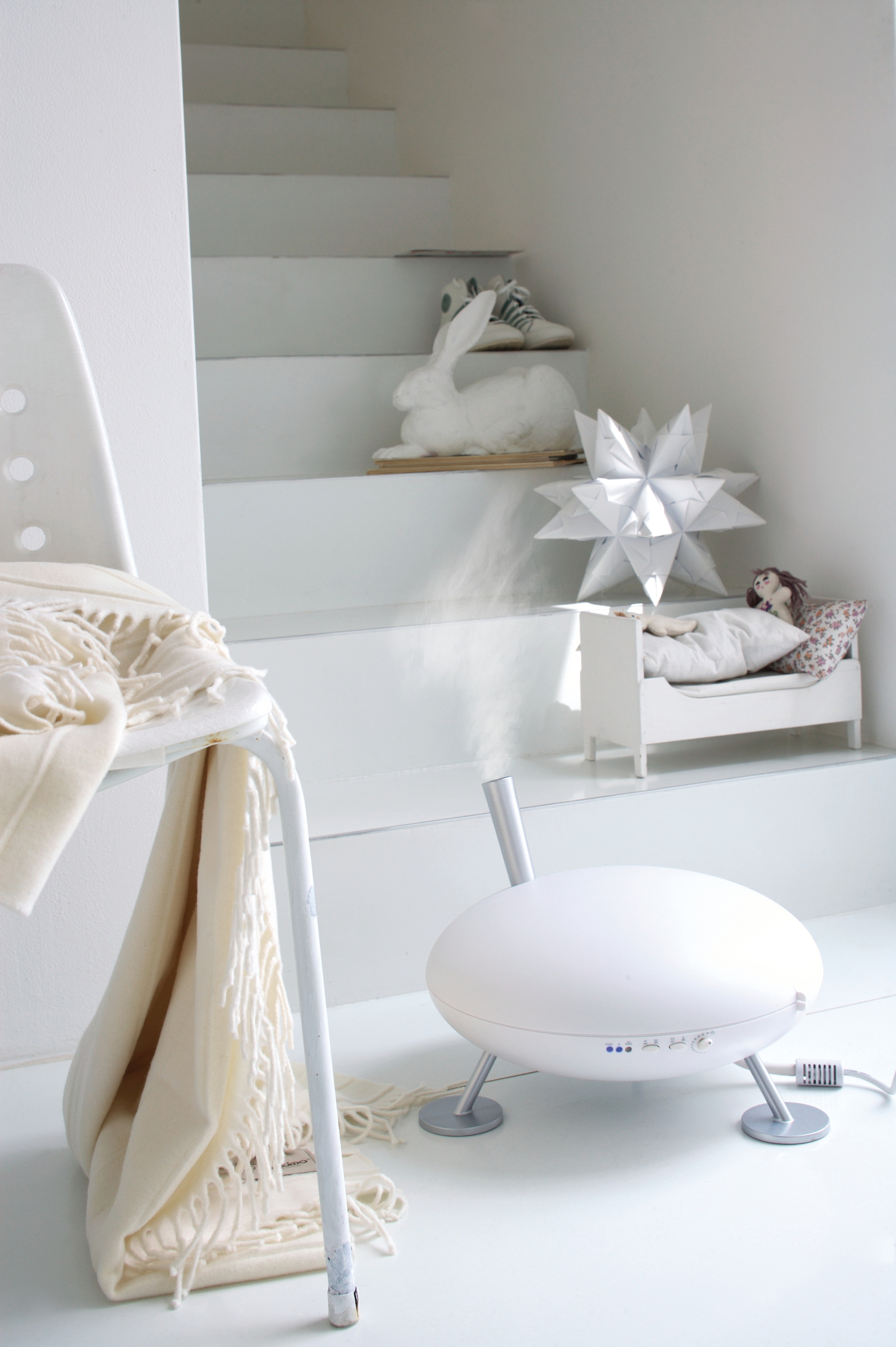 Ein weißes Zimmer mit Stuhl, Decke und Luftbefeuchter. Eine Puppe liegt in einem Bett auf einer Treppe.