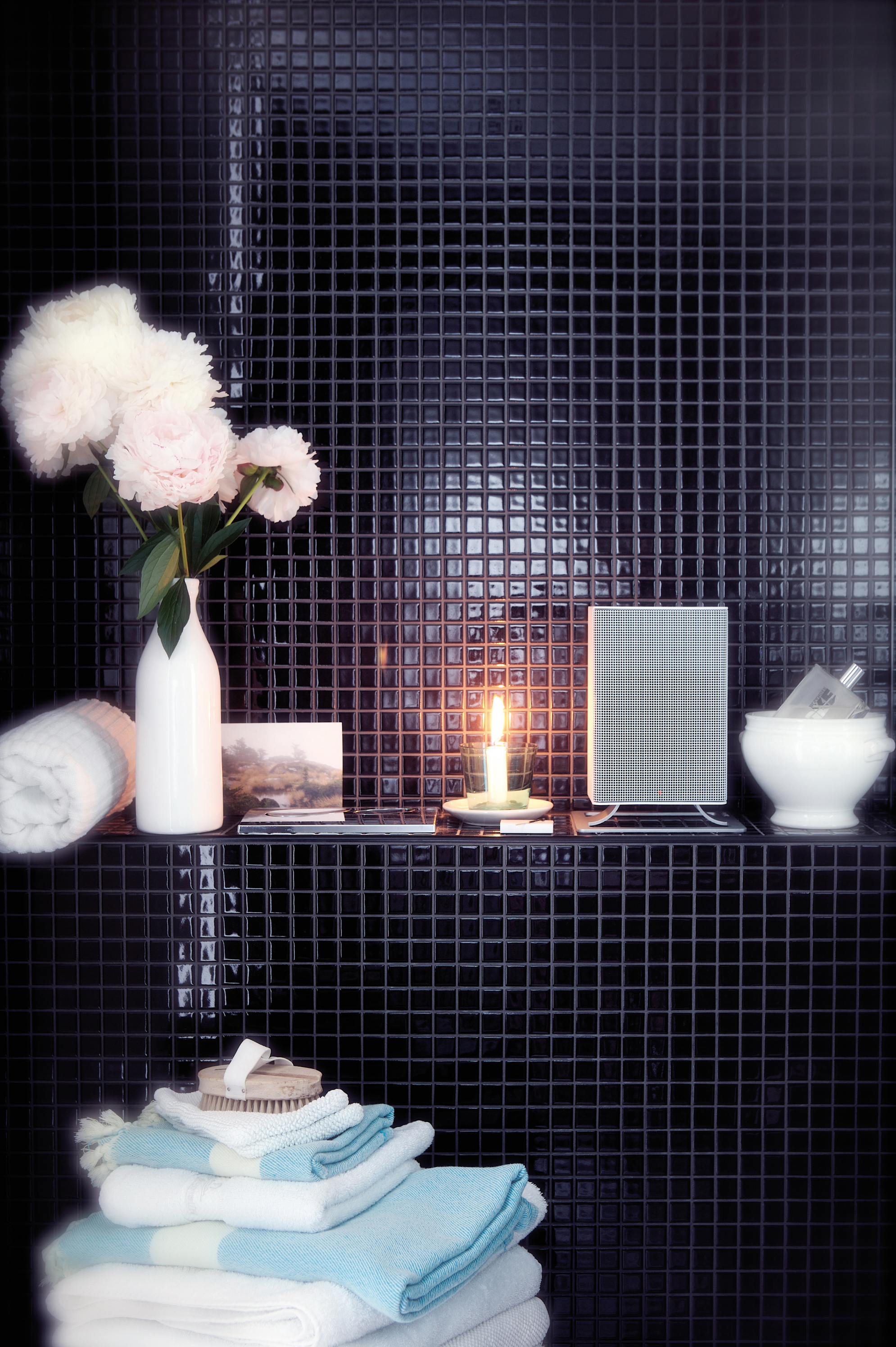 Ein Badezimmerregal mit einer weißen Blumenvase, einer Kerze, einem Lautsprecher und Handtüchern.
