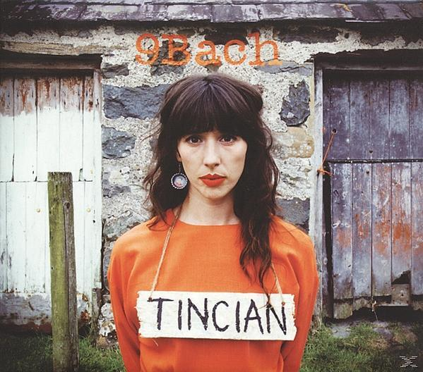 Nő narancssárga ingben, 'TINCIA' felirattal, kőfal és ajtók előtt. A feje fölött '9Bach' felirat olvasható.