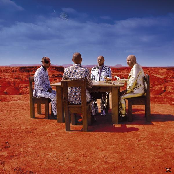 Vier Männer an einem Tisch in Wüstenlandschaft. Rote Erde und blauer Himmel.