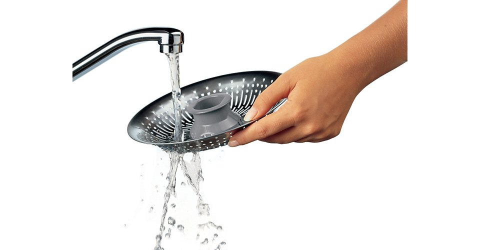 Main tenant une passoire argentée sous l'eau courante, nettoyant un insert de passoire grise.
