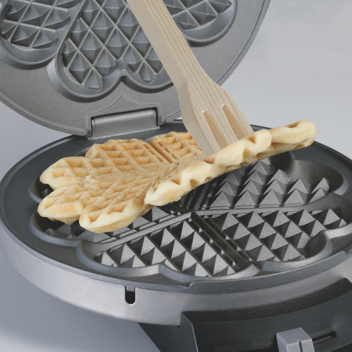 Waffeleisen mit einem herzförmigen Waffel, die mit einer Holzgabel herausgehoben wird. Heller Hintergrund. Die Waffel ist goldbraun.
