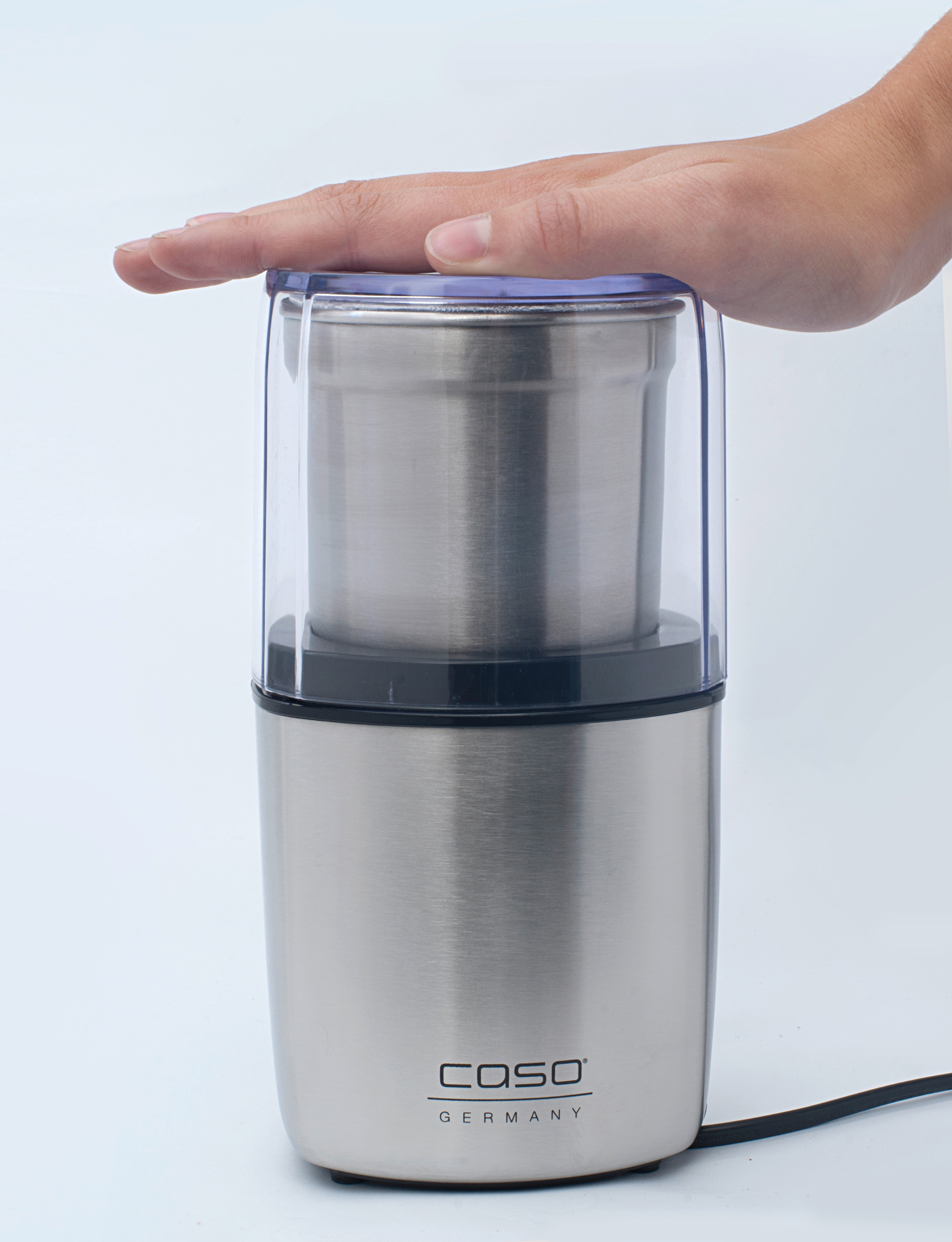 Eine Hand, die einen transparenten Deckel auf eine silberne Kaffeemühle setzt.