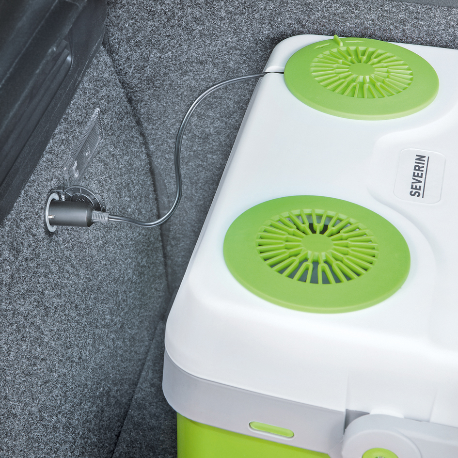 Eine weiße und grüne Kühlbox mit Stromkabel, das in einen grauen Autoinnenraum gesteckt ist.
