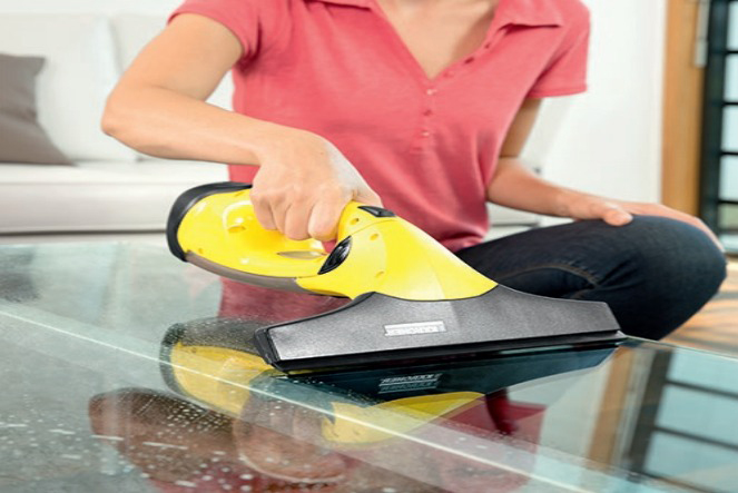Een persoon die een glazen oppervlak schoonmaakt met een gele handmatige glasreiniger. Reflectie van de reiniger zichtbaar.