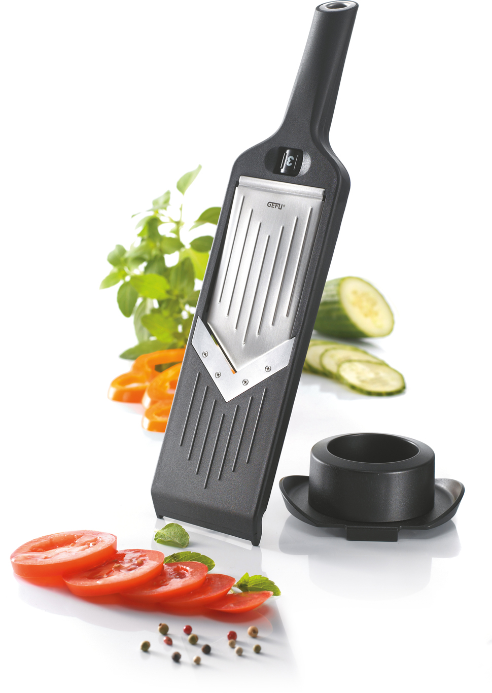 Ein schwarzer Lebensmittelschneider mit Tomatenscheiben, Basilikum und Pfefferkörnern auf weißer Oberfläche.