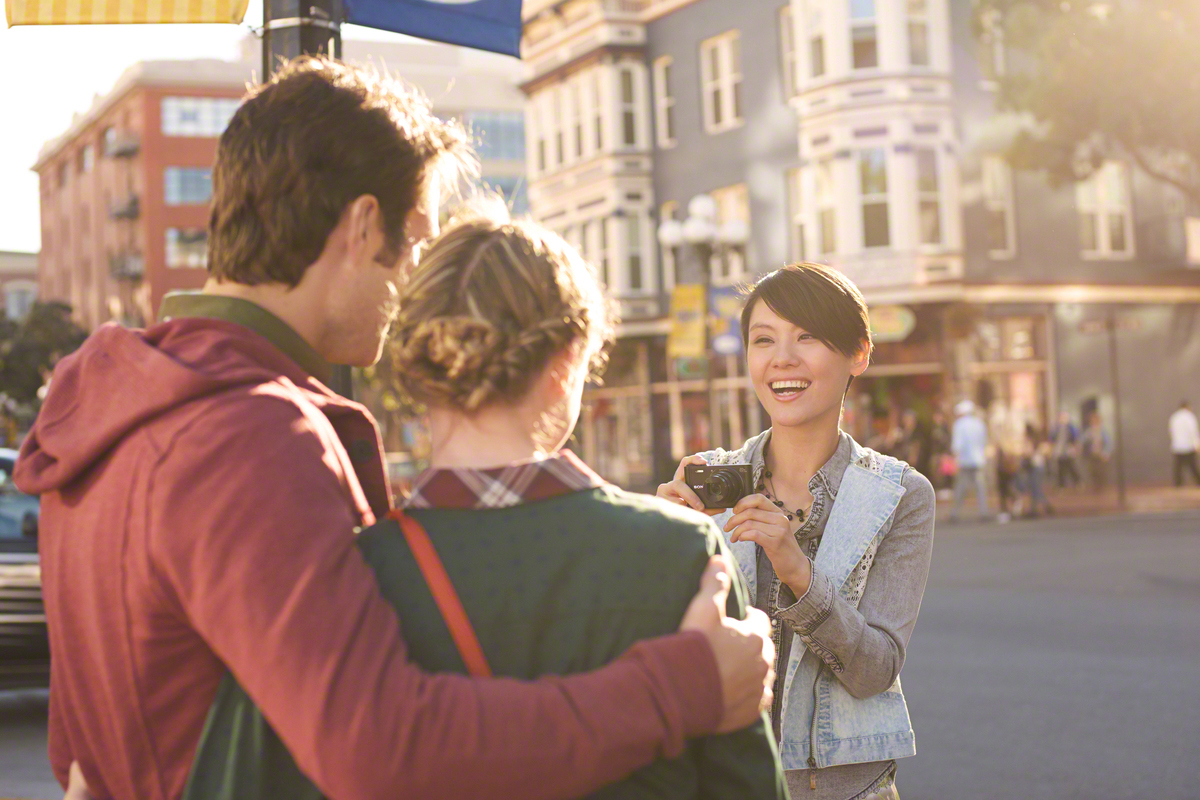 Eine Frau fotografiert ein sich umarmendes Paar auf einer Stadtstraße. Gebäude im Hintergrund.