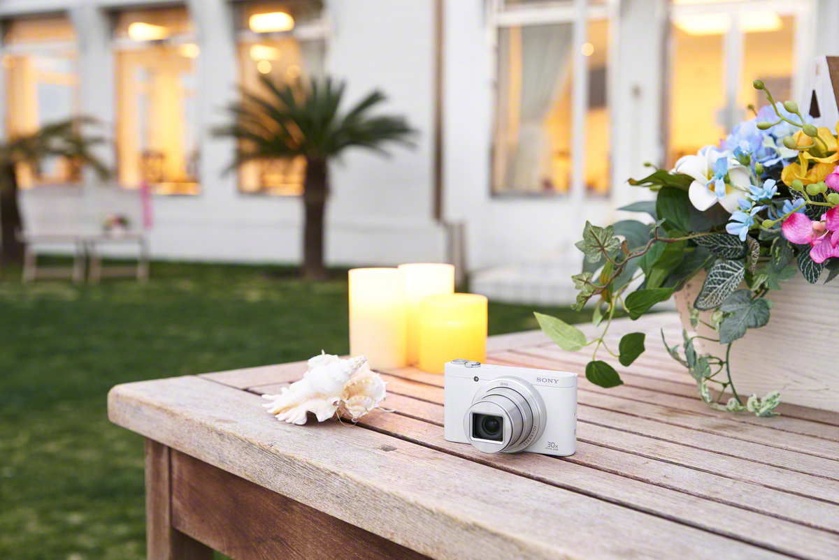 Weiße Sony-Kamera, Kerzen, Muschel und Blumen auf einem Holztisch mit Gebäude und grünem Gras im Hintergrund.