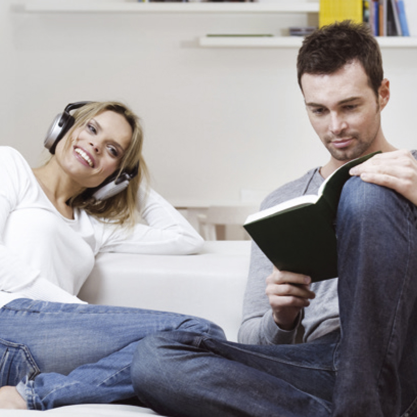 A woman with headphones smiles while a man reads a book, both on a white couch.