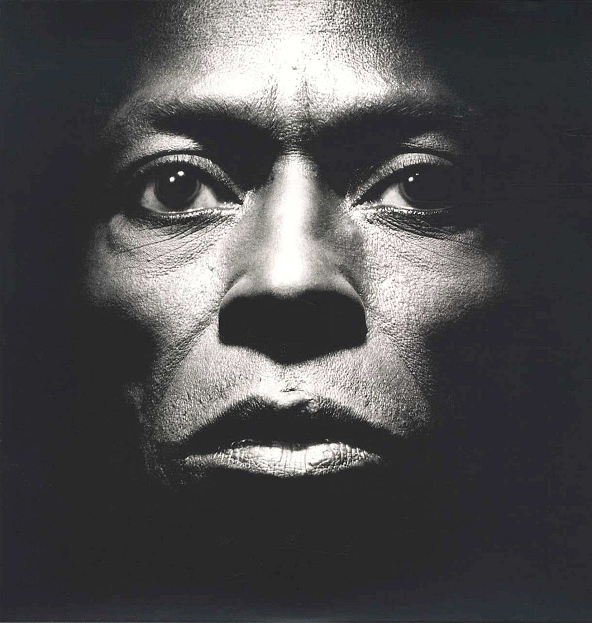 Schwarz-Weiß-Foto von Miles Davis' Gesicht. Nahaufnahme, intensiver Blick, schwarzer Hintergrund.
