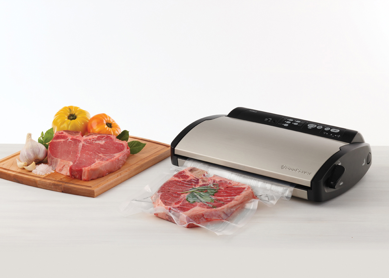Raw steaks on a wooden board, a vacuum sealer with a steak in a bag, and vegetables.