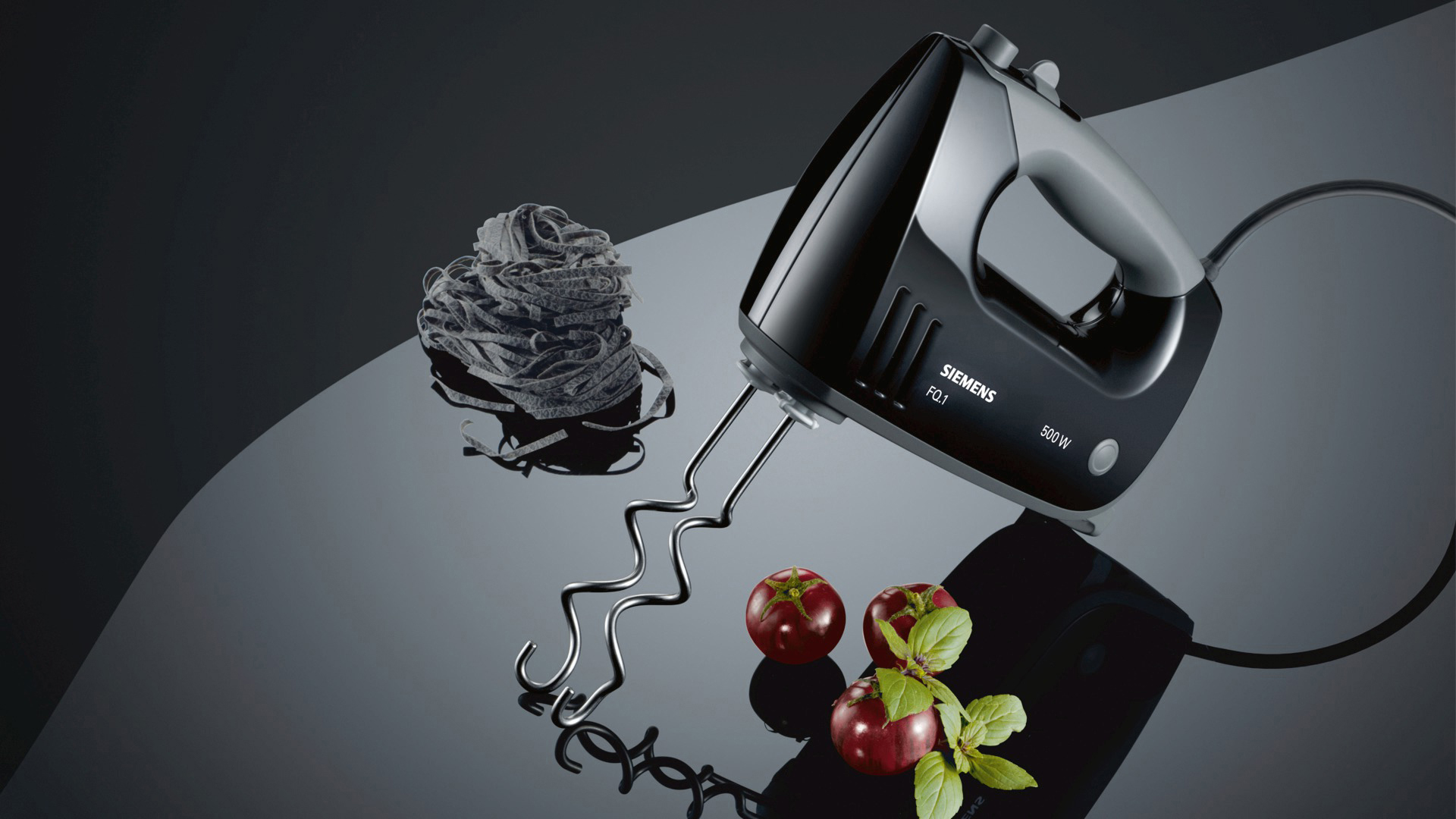 Ein schwarzer Handmixer mit schwarzer Pasta, roten Tomaten und Basilikumblättern auf einer reflektierenden Oberfläche.