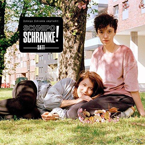 Zwei Frauen auf Gras, eine liegend, eine sitzend, ein Baum und ein Gebäude.