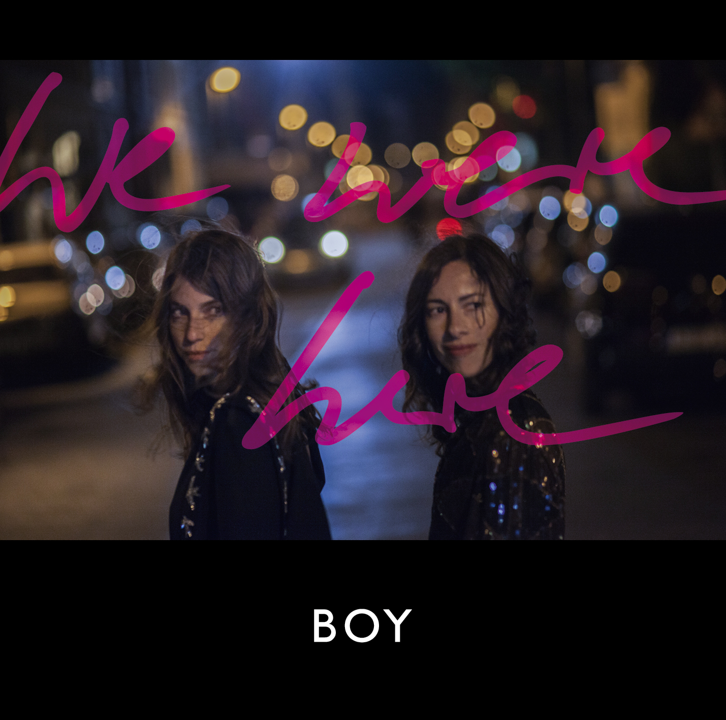 Albumcover mit zwei Frauen vor einer verschwommenen Straßenszene. Rosa 'We Are Here' Text.