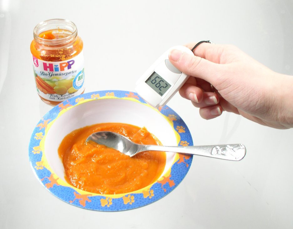 Löffel in Schüssel mit Babynahrung. Gläschen Babynahrung und Hand hält Gerät.