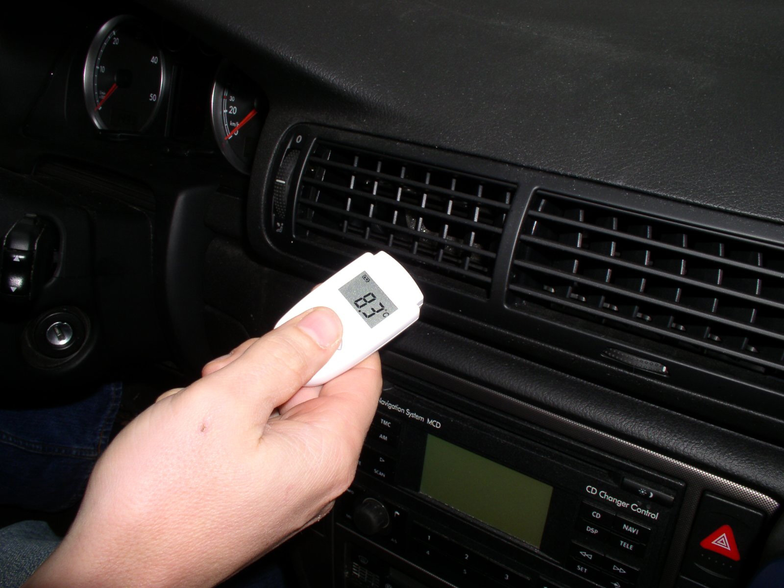 Hand, die ein Thermometer im Auto hält, zeigt 83,3 an. Lüftungsgitter und Armaturenbrett sind im Hintergrund sichtbar.