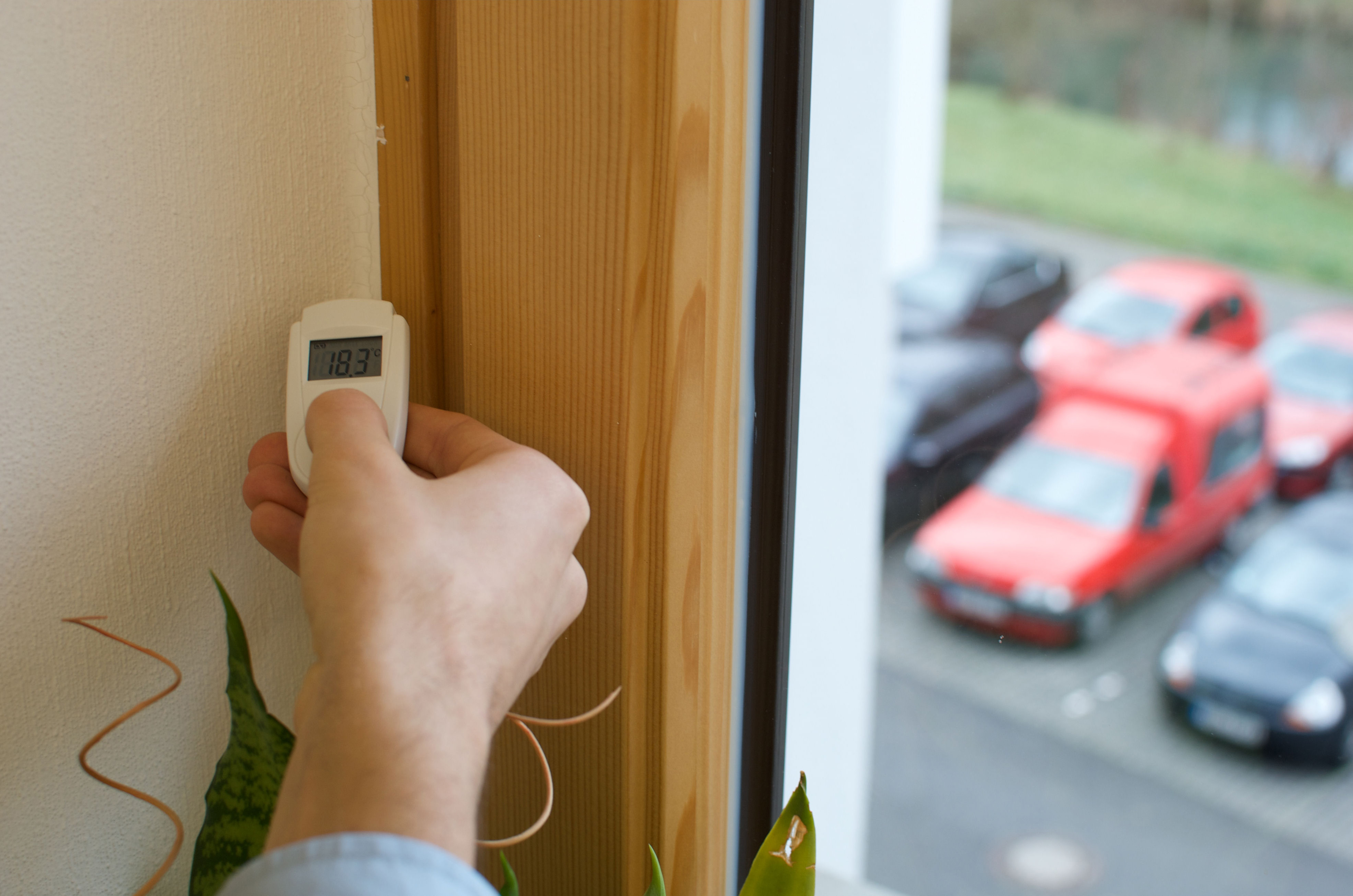 Eine Hand, die ein weißes Gerät mit der Anzeige 18,3 in der Nähe eines Fensters hält, mit Autos draußen.