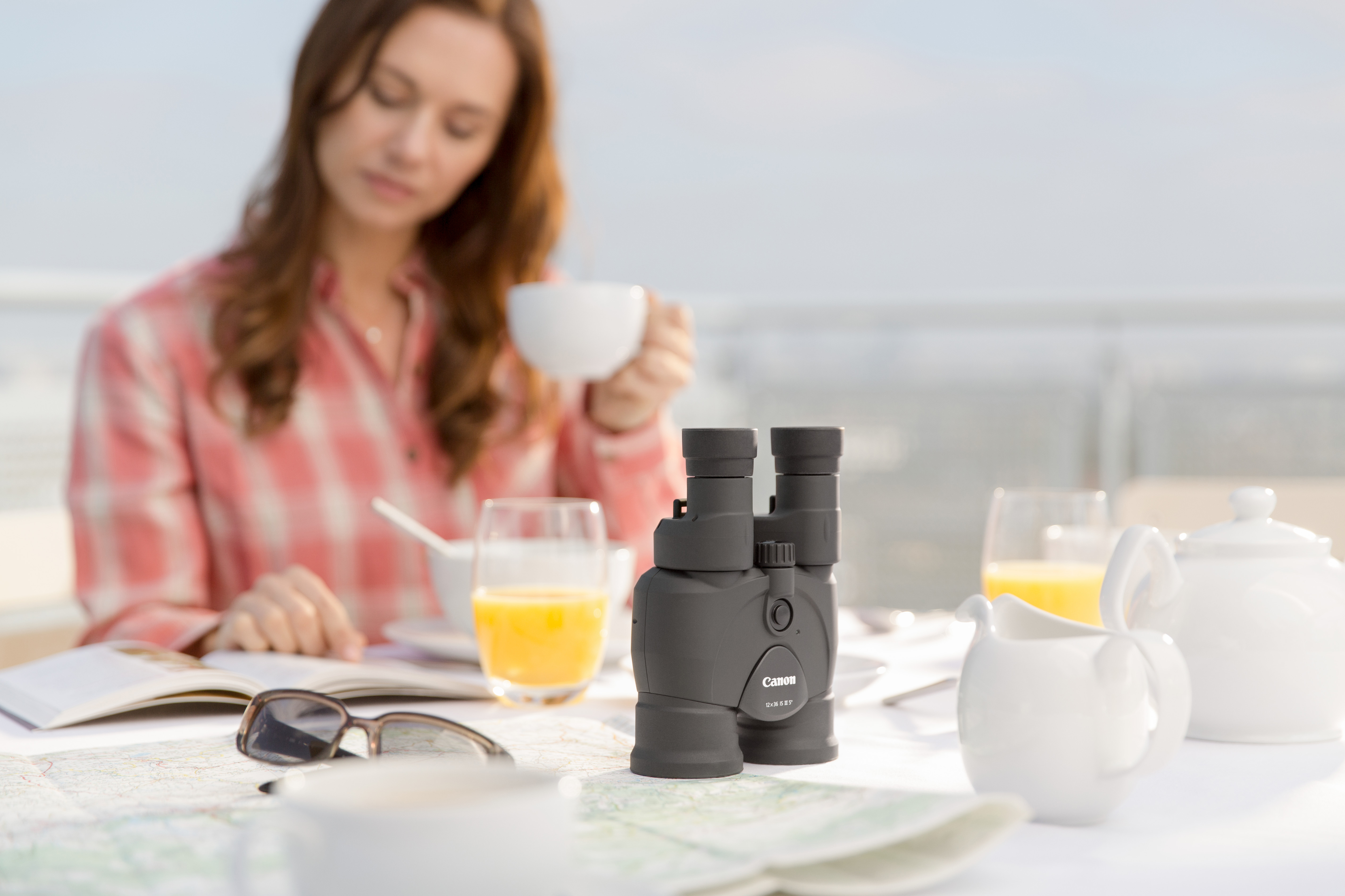Donna a un tavolo con binocolo, tazza di caffè e un libro. Sono presenti anche succo d'arancia e una teiera.