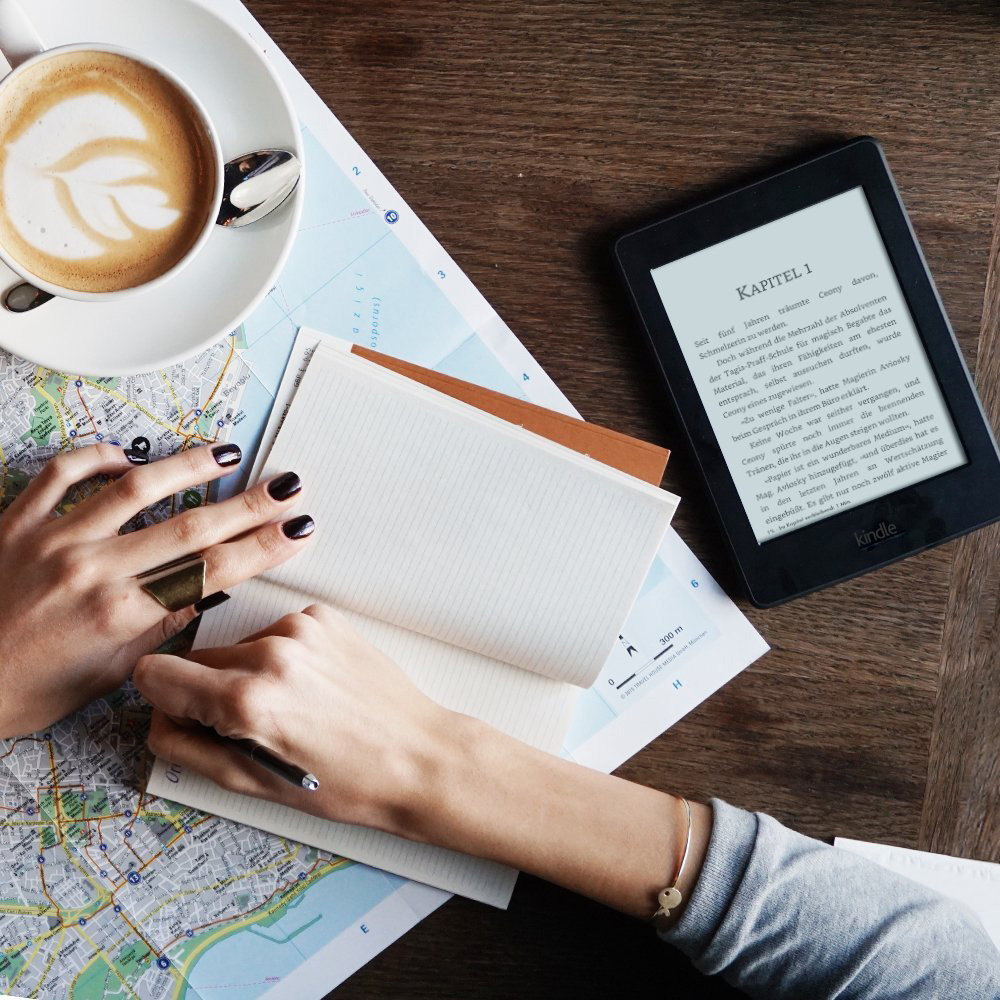 Hände einer Person, die in ein Notizbuch schreiben. Es gibt eine Tasse Kaffee und ein Kindle.