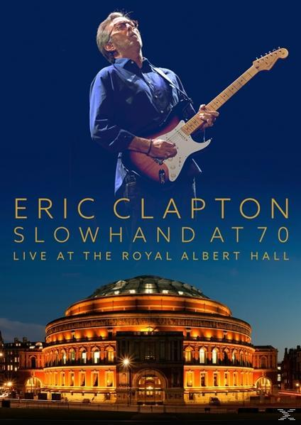 Eric Clapton spielt Gitarre auf der Bühne, mit einem großen Gebäude im Hintergrund.