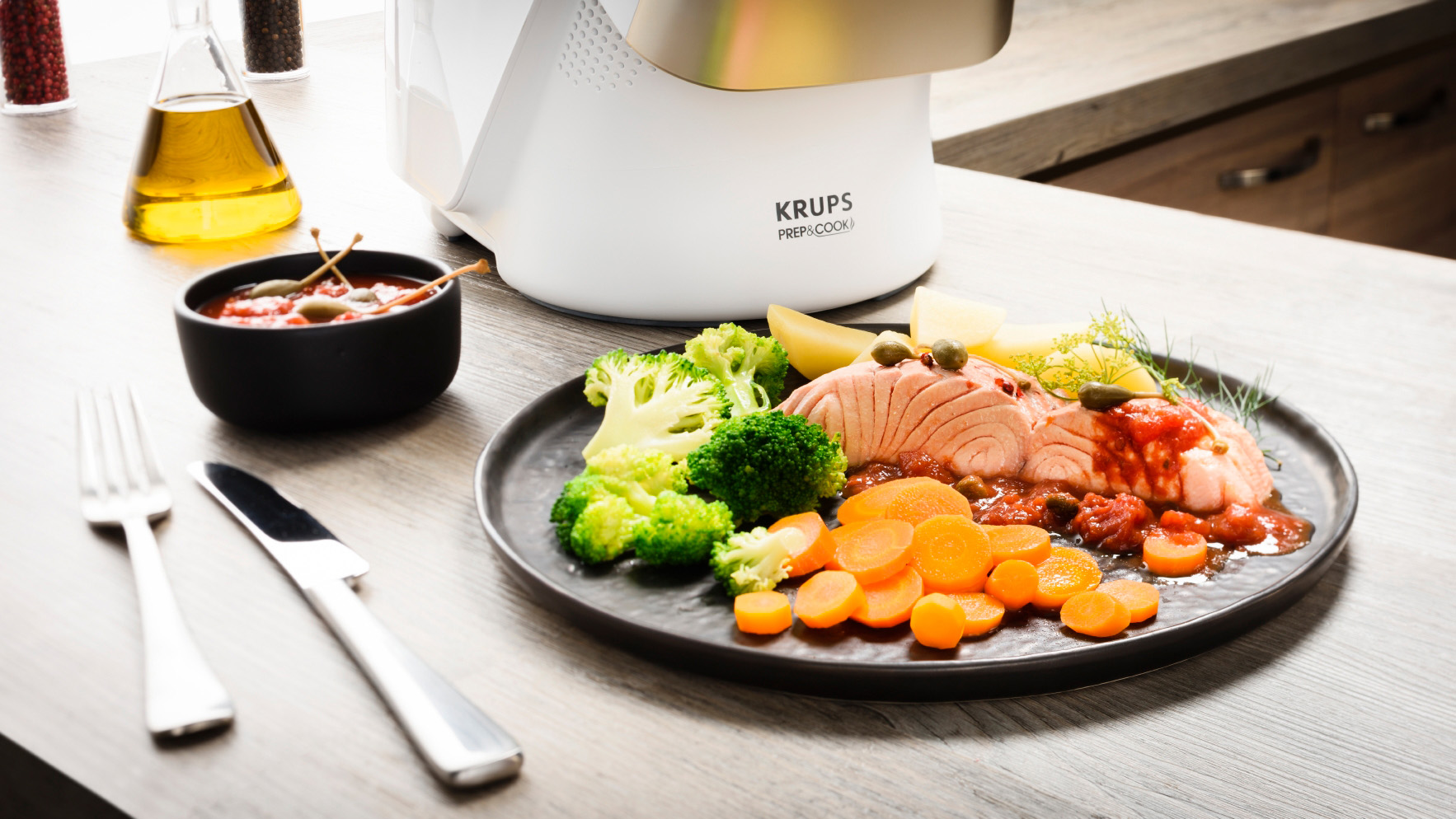 Lachsgericht mit Gemüse und einem weißen Foodprozessor. Das Essen ist auf einem schwarzen Teller.