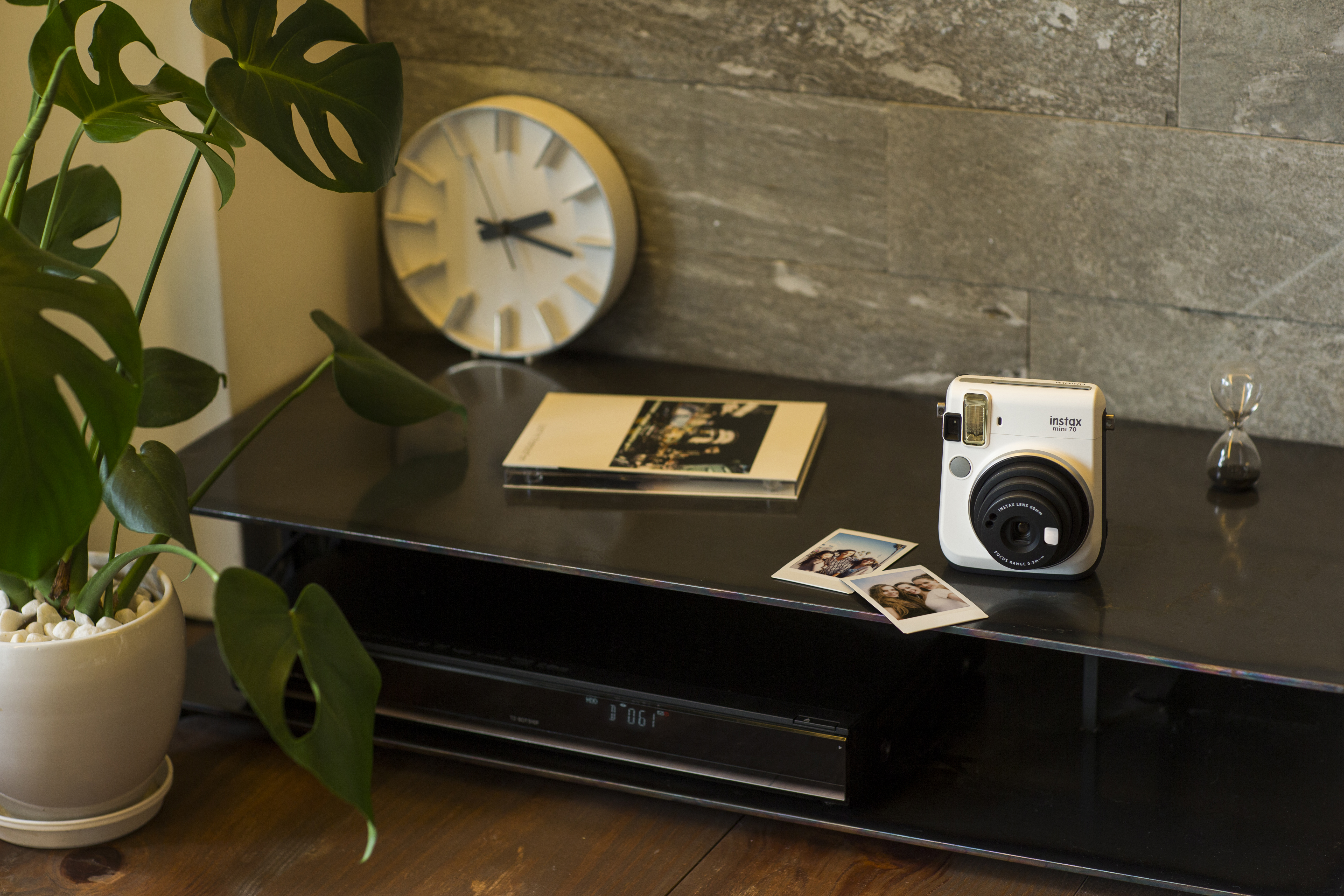 Ein schwarzes Regal mit einer weißen Instax-Kamera, einer Uhr, einer Pflanze und Fotos. Eine kleine Sanduhr ist ebenfalls vorhanden.