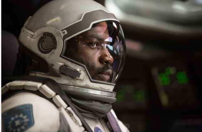 Astronaute en combinaison blanche, à l'intérieur d'un vaisseau spatial. Le casque reflète l'intérieur. Le fond est sombre.