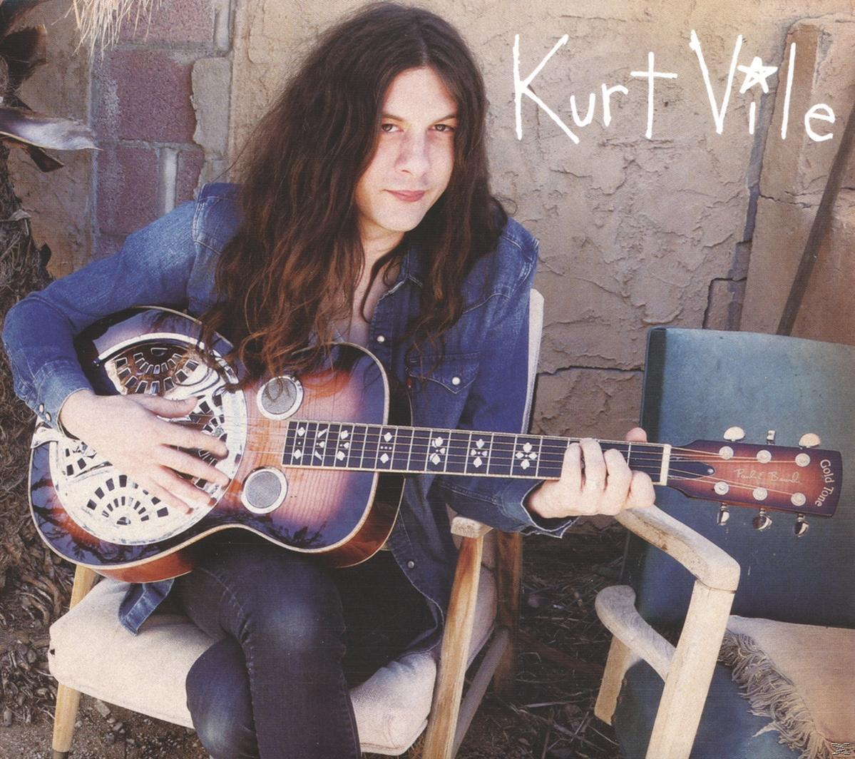Kurt Vile spielt Gitarre, sitzt auf einem Stuhl, trägt eine Jeansjacke. Eine Backsteinmauer ist der Hintergrund.