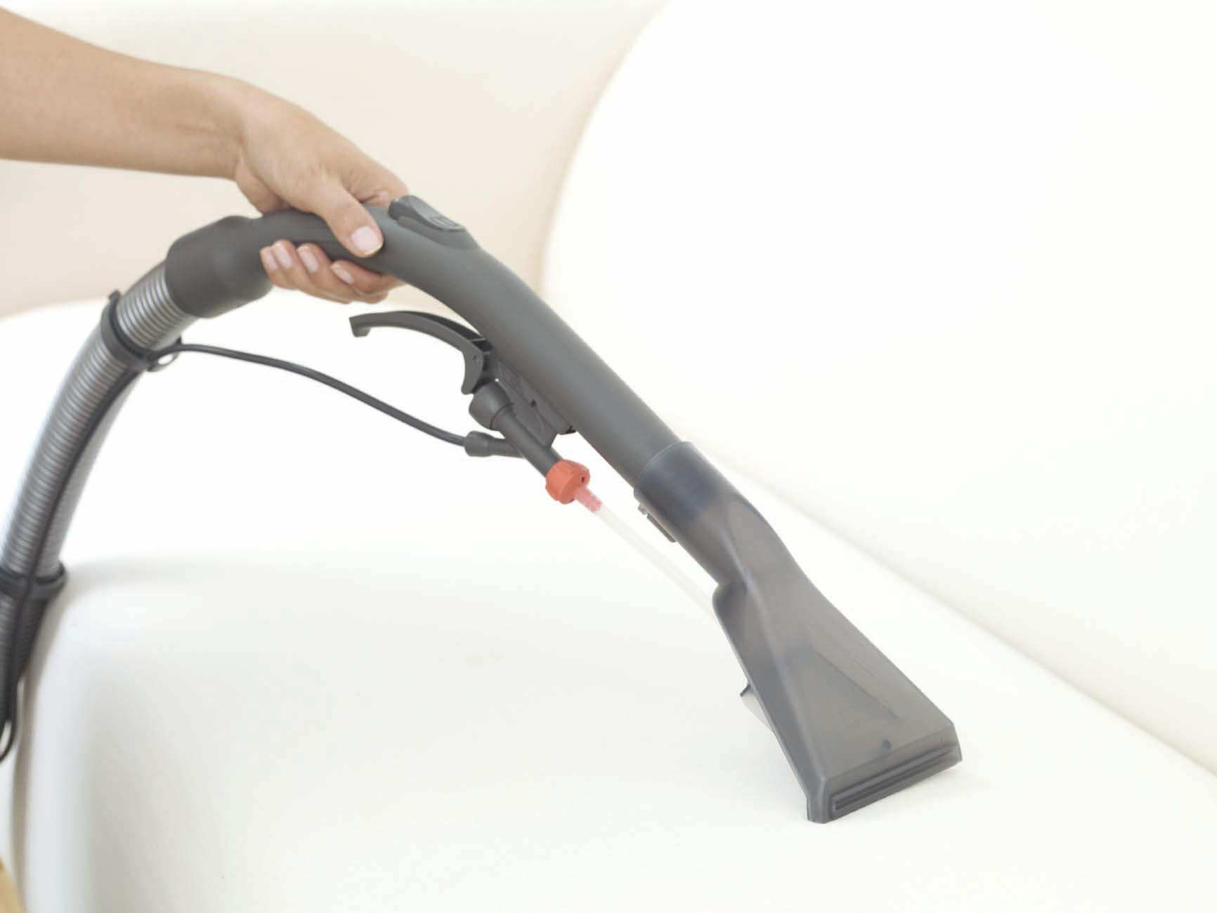 Eine Hand, die ein weißes Sofa mit einem grauen Staubsaugeraufsatz absaugt.