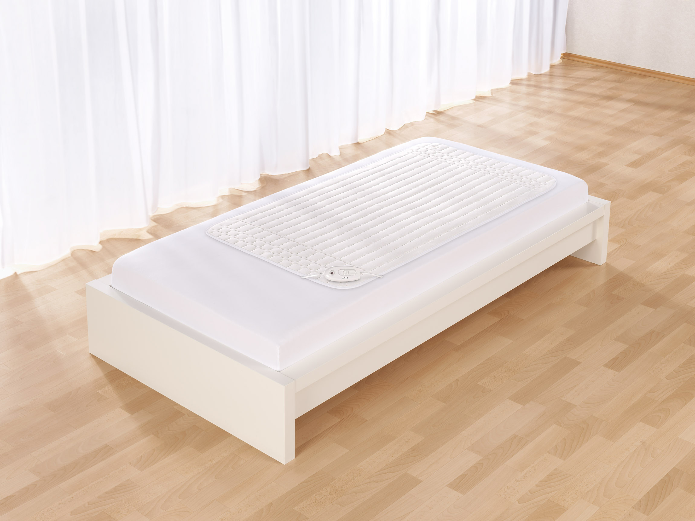 Weißes Bett mit Heizkissen, vor weißem Vorhang und Holzboden.