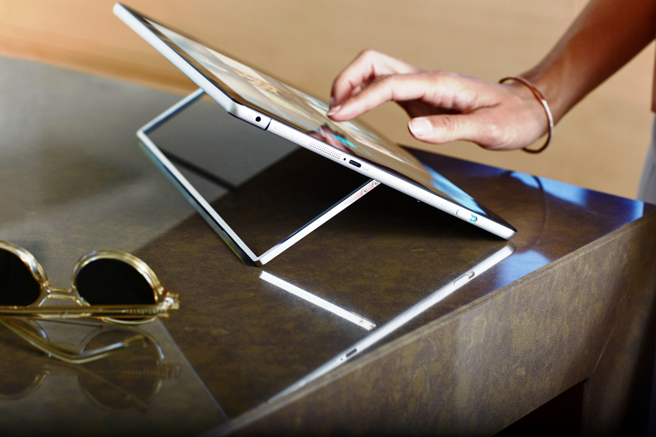 Eine Hand tippt auf ein Tablet auf einem Tisch, daneben liegen goldene Sonnenbrillen.