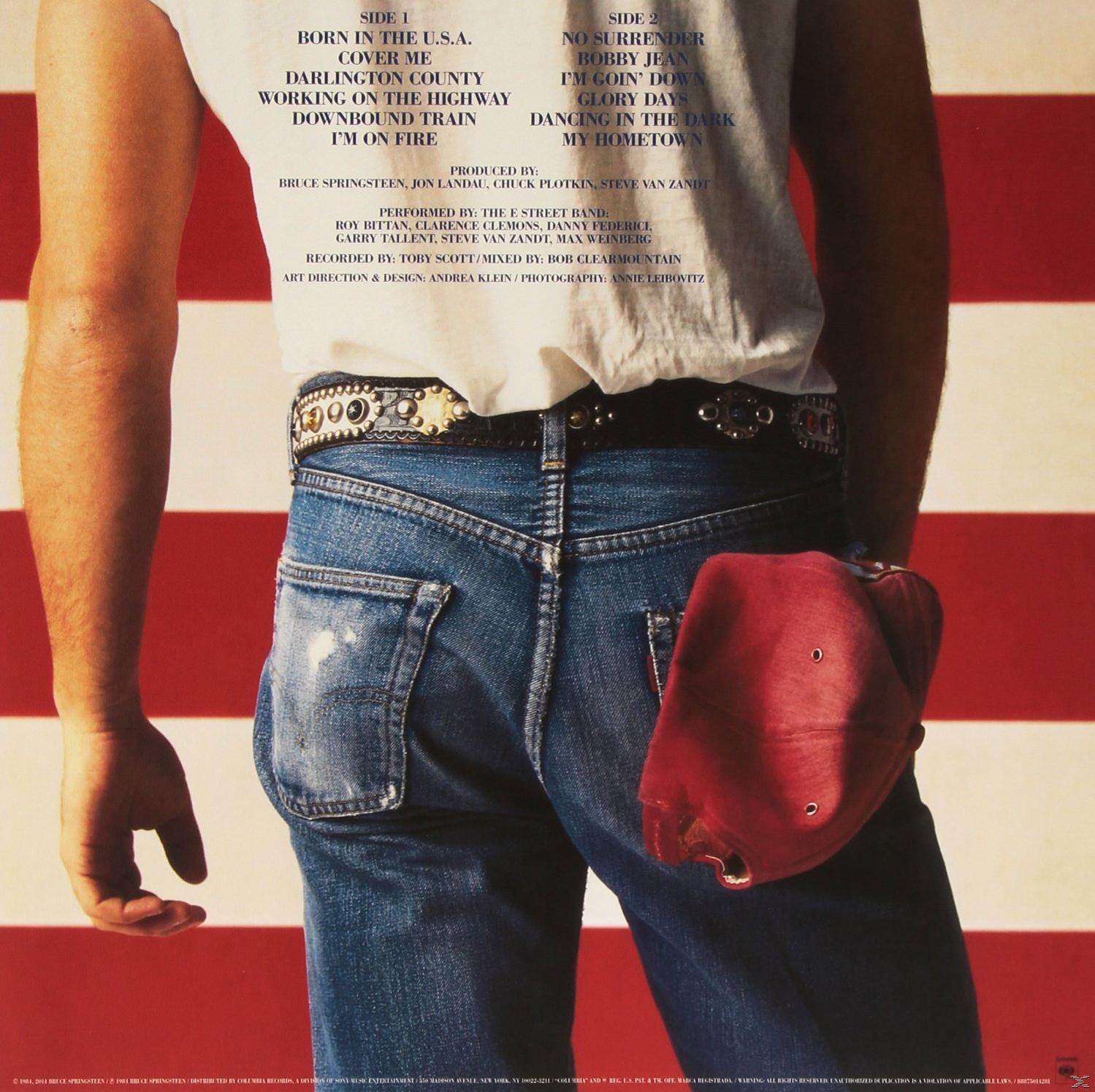 Rückseite eines Mannes mit Blue Jeans, einem Gürtel mit Nieten und einer roten Kappe. Im Hintergrund eine US-Flagge.