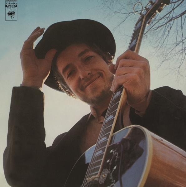 Bob Dylan hält eine Gitarre, lächelt, vor blauem Himmel.