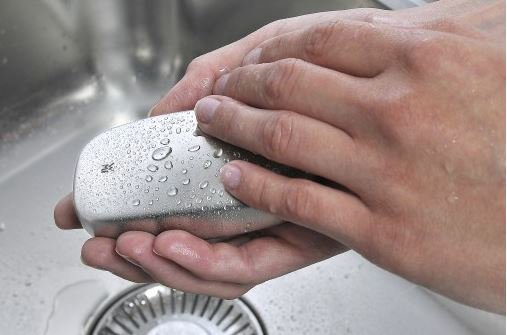Hände, die einen Metallseifenriegel unter fließendem Wasser in einem Edelstahlbecken waschen. Wassertröpfchen sind sichtbar.