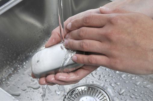 Hände waschen einen silbernen Gegenstand unter fließendem Wasser in einem Edelstahlbecken. Das Wasser ist klar.