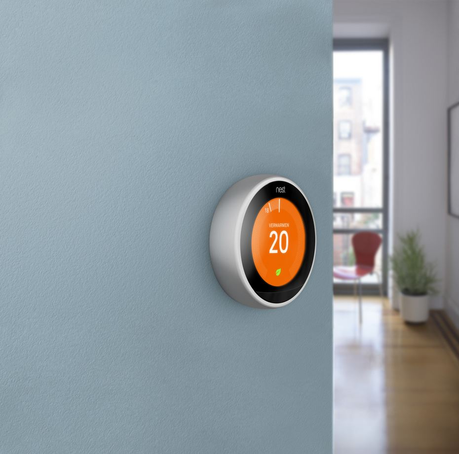 Een slimme thermostaat gemonteerd op een blauwe muur, met een temperatuur van 20 graden. Een kamer staat op de achtergrond.