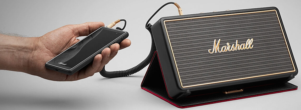Une main tenant un téléphone connecté à un haut-parleur Marshall, avec une finition noire et dorée.