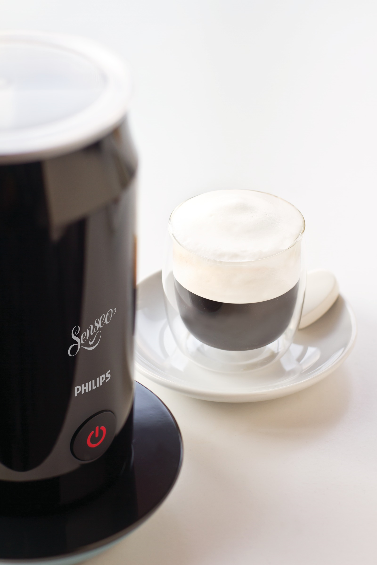 Een koffiezetapparaat, een kopje koffie met schuim en een schotel. De achtergrond is wit.