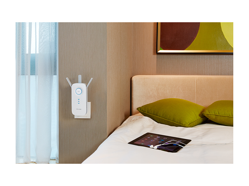 A white WiFi extender is plugged into a wall socket. A tablet and headphones are on a bed next to it.