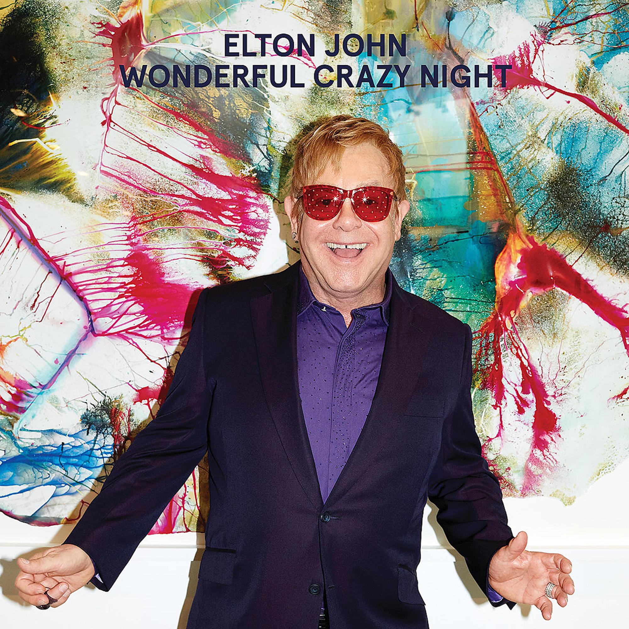 Elton John trägt Sonnenbrille, lächelt, vor farbenfrohem Hintergrund.