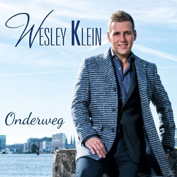 Man in een geruite jas, glimlachend, met stad en water op de achtergrond. Tekst luidt 'Wesley Klein' en 'Onderweg'.