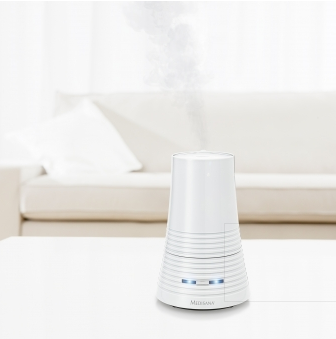 Humidificateur d'air blanc sur une table blanche, libérant de la vapeur, avec un canapé blanc en arrière-plan flou.