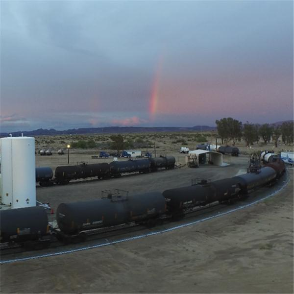 Een trein met zwarte tankwagens staat op rails. Een regenboog spant zich uit over een donkere hemel boven een woestijnlandschap.