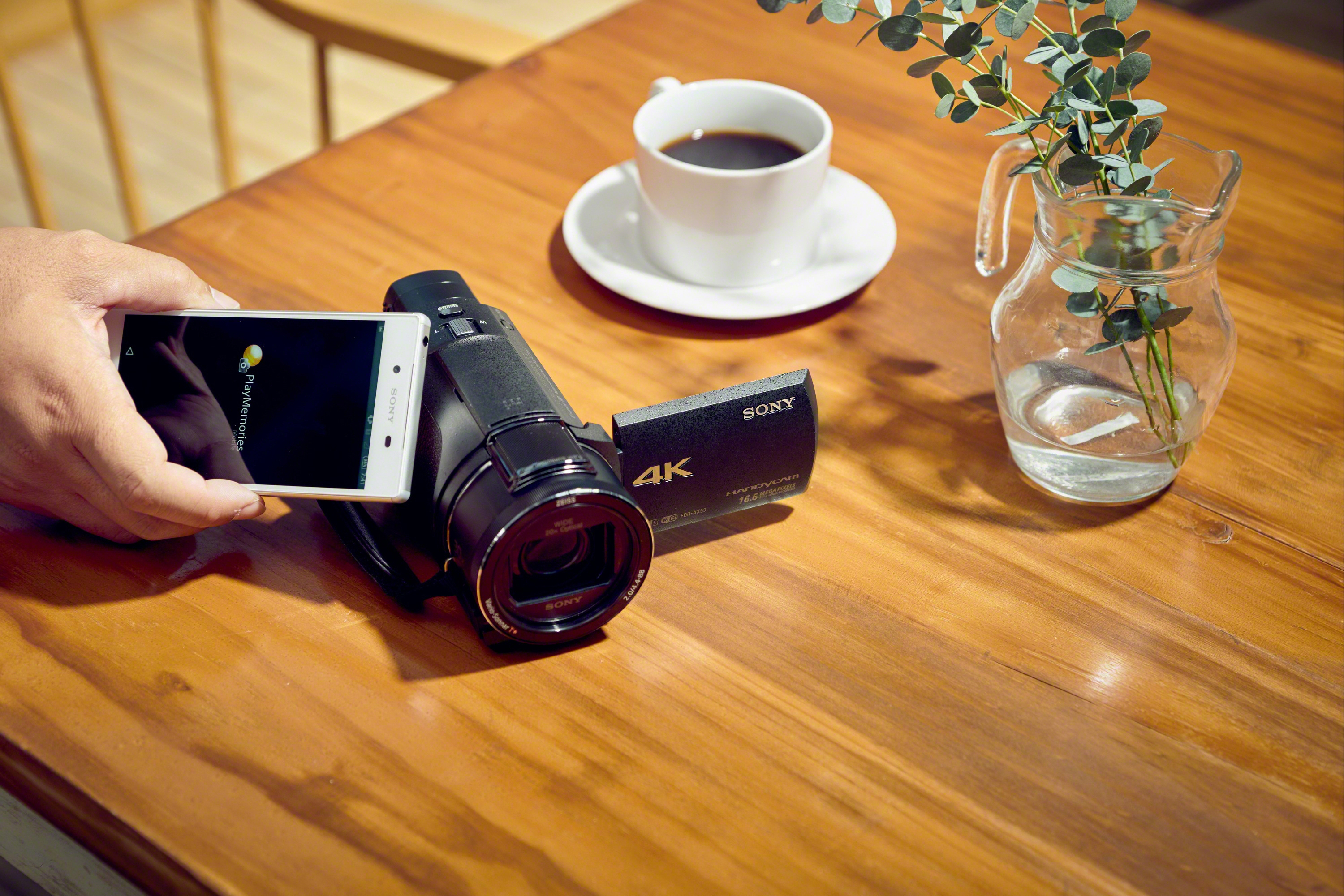 Una persona che tiene un telefono che mostra una fotocamera, una fotocamera con una tazza di caffè e fiori.