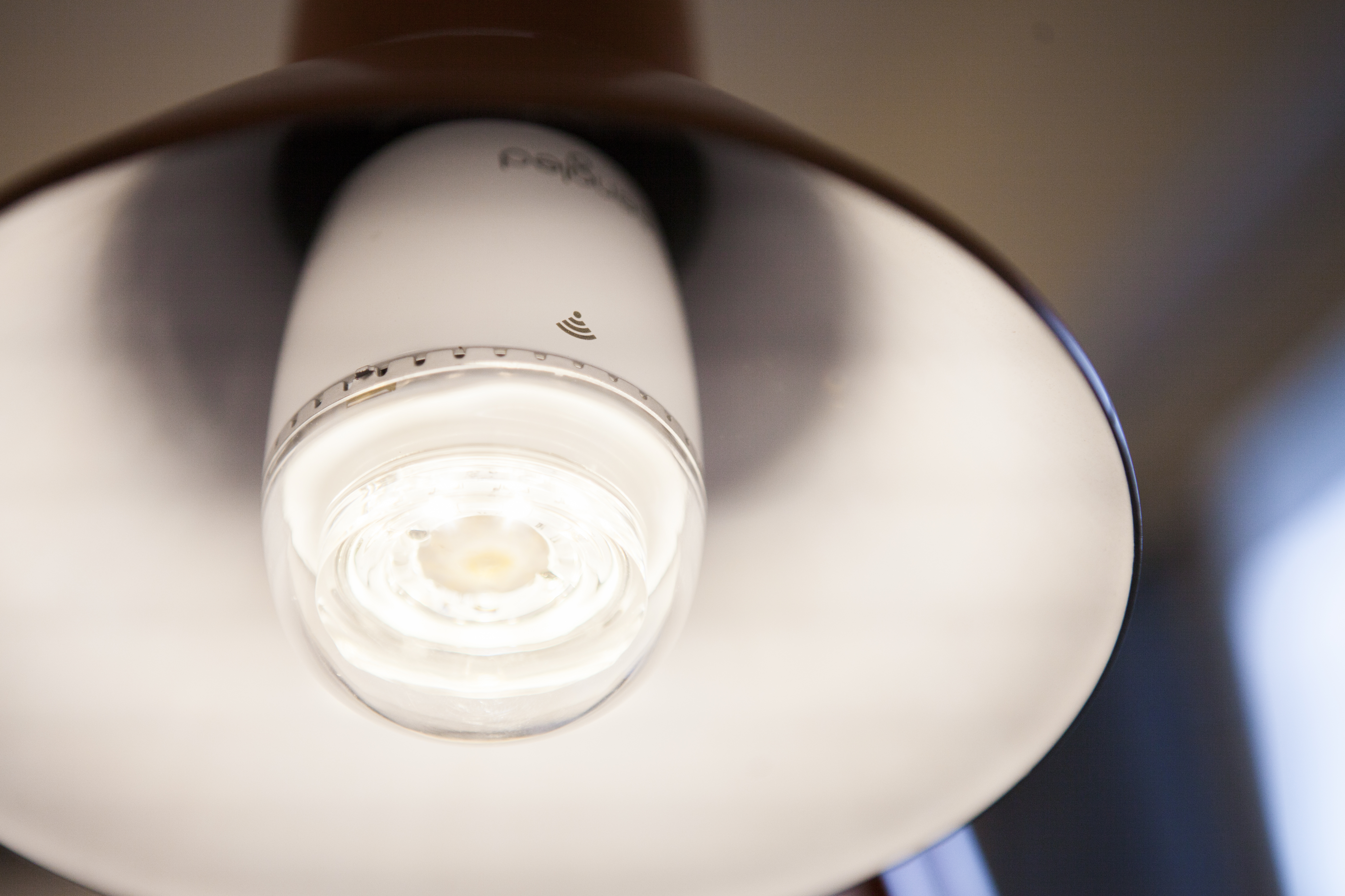 A white smart bulb is lit inside a lamp. The lamp shade is brown and white. The background is blurred.
