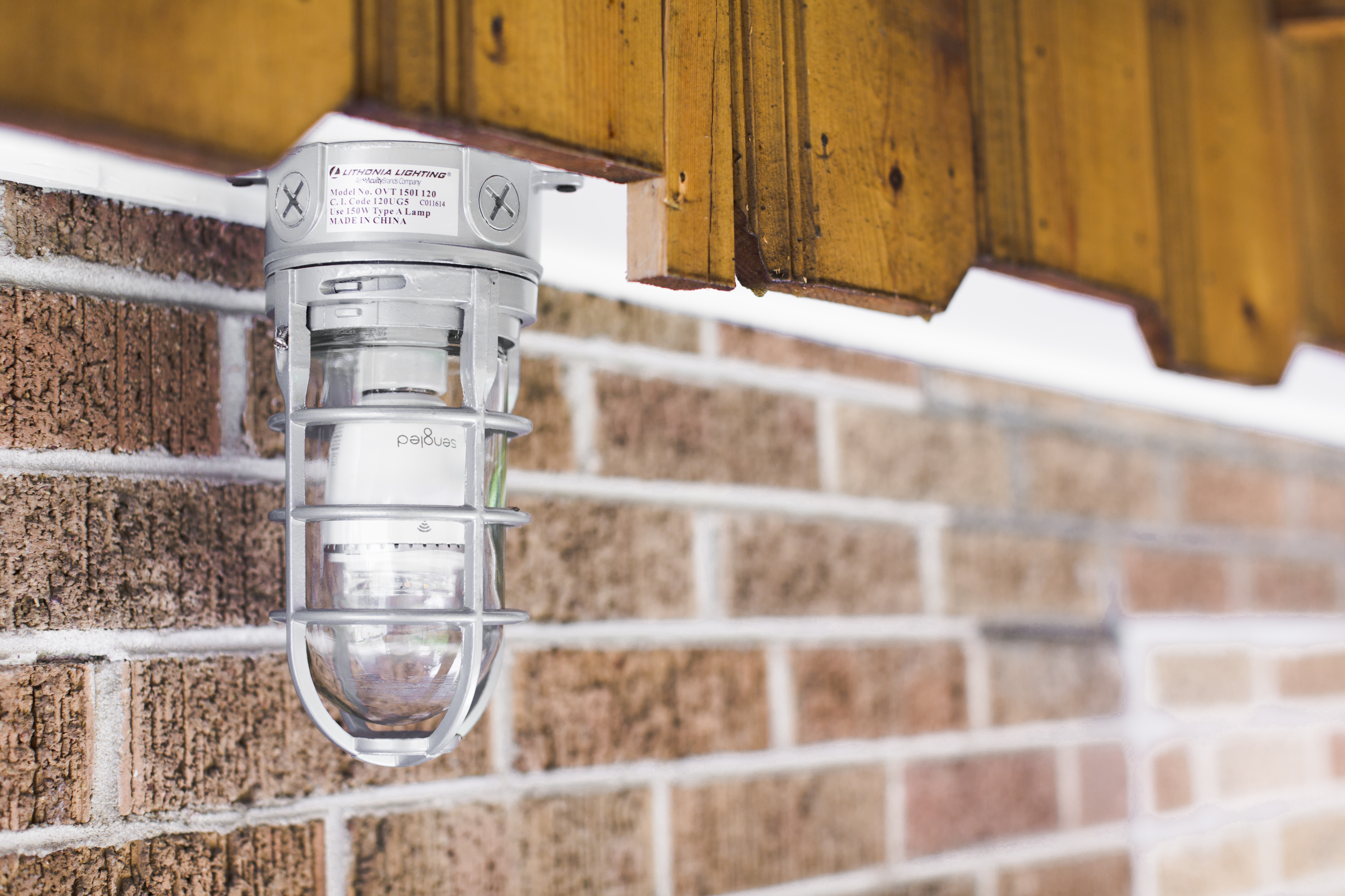 A vintage-style light fixture is mounted on a brick wall under a wooden overhang. The light has a metal cage and glass.
