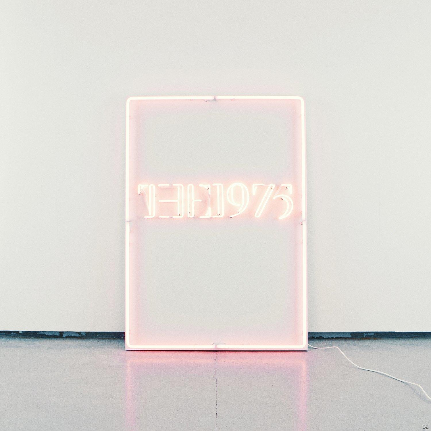Neonschild mit dem Bandnamen "THE 1975" in einem rosa Rechteck an einer weißen Wand.