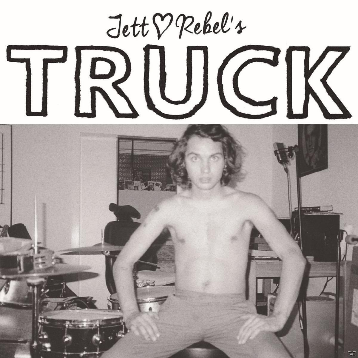 Een man zonder shirt zit met zijn handen op zijn knieën. Drums en een microfoon staan achter hem. Het woord "TRUCK" staat boven zijn hoofd.